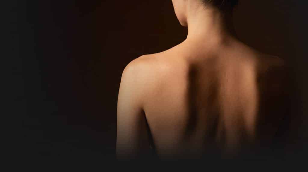 Woman's bare back and shoulder photographed from behind in warm dramatic lighting against a dark background